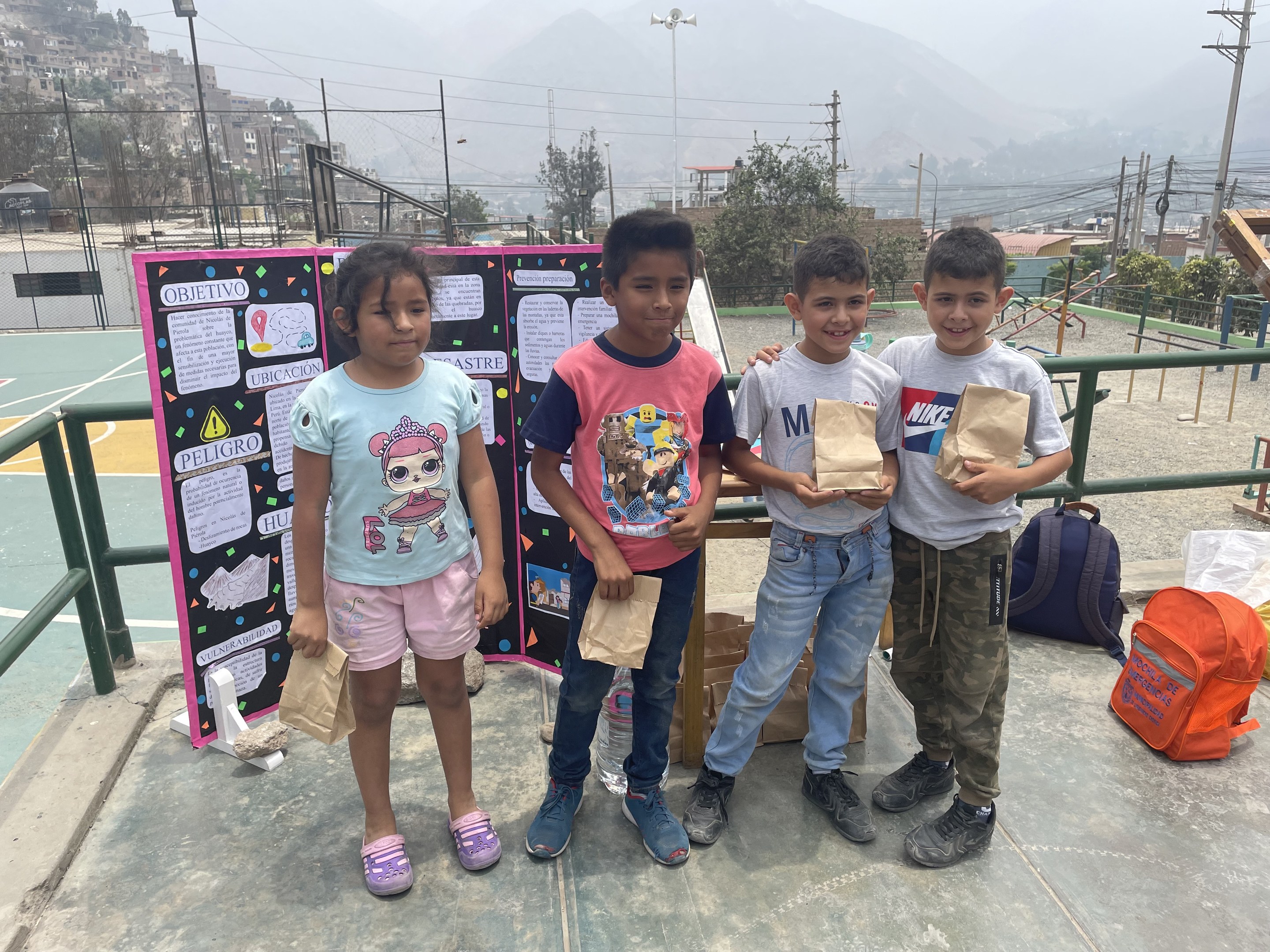 Gruppe von Kindern auf einem Basketballfeld mit Papiertüten in den H├Ąnden, mit Tüten und Gegenst├Ąnden rechts und einer Tafel mit Text links, vor einer ├Ąu├čeren Kulisse mit Zaun, Masten, Stra├čenlaternen, B├Ąumen, Geb├Ąuden, H├Ąngen und Himmel.