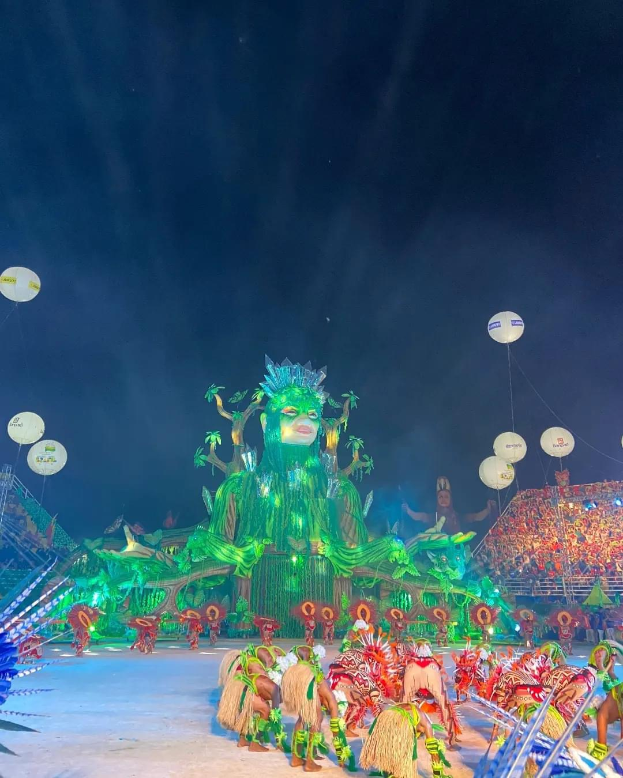 Ein lebendiger Karneval in Rio de Janeiro, Brasilien, mit Performern in farbenfrohen Kostümen auf der Bühne, einer großen Menge im Publikum und einem Himmel voller bunter Lichter und Ballons.