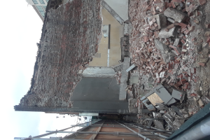 Teilweise abgerissenes Gebäude mit verstreuten Ziegeln und Metallstangen, andere Gebäude und einen klaren blauen Himmel im Hintergrund.