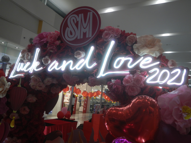 Ein Geschäft mit einem Schild, das "Glück und Liebe 2021" trägt, umgeben von Blumen, Ballons und anderen Dekorationen, mit anderen Geschäften, Lichtern und einer Wand im Hintergrund, die eine festliche Atmosphäre für die SM-Valentinstag-Feier schaffen.