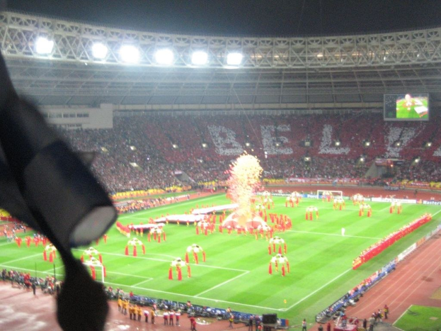 Ein Stadion mit Flutlicht, Eisengittern, Zuschauern, stehenden Personen, Werbetafeln, Anzeigetafeln, Sicherheitskamera, Flaggen und Dekorationen.