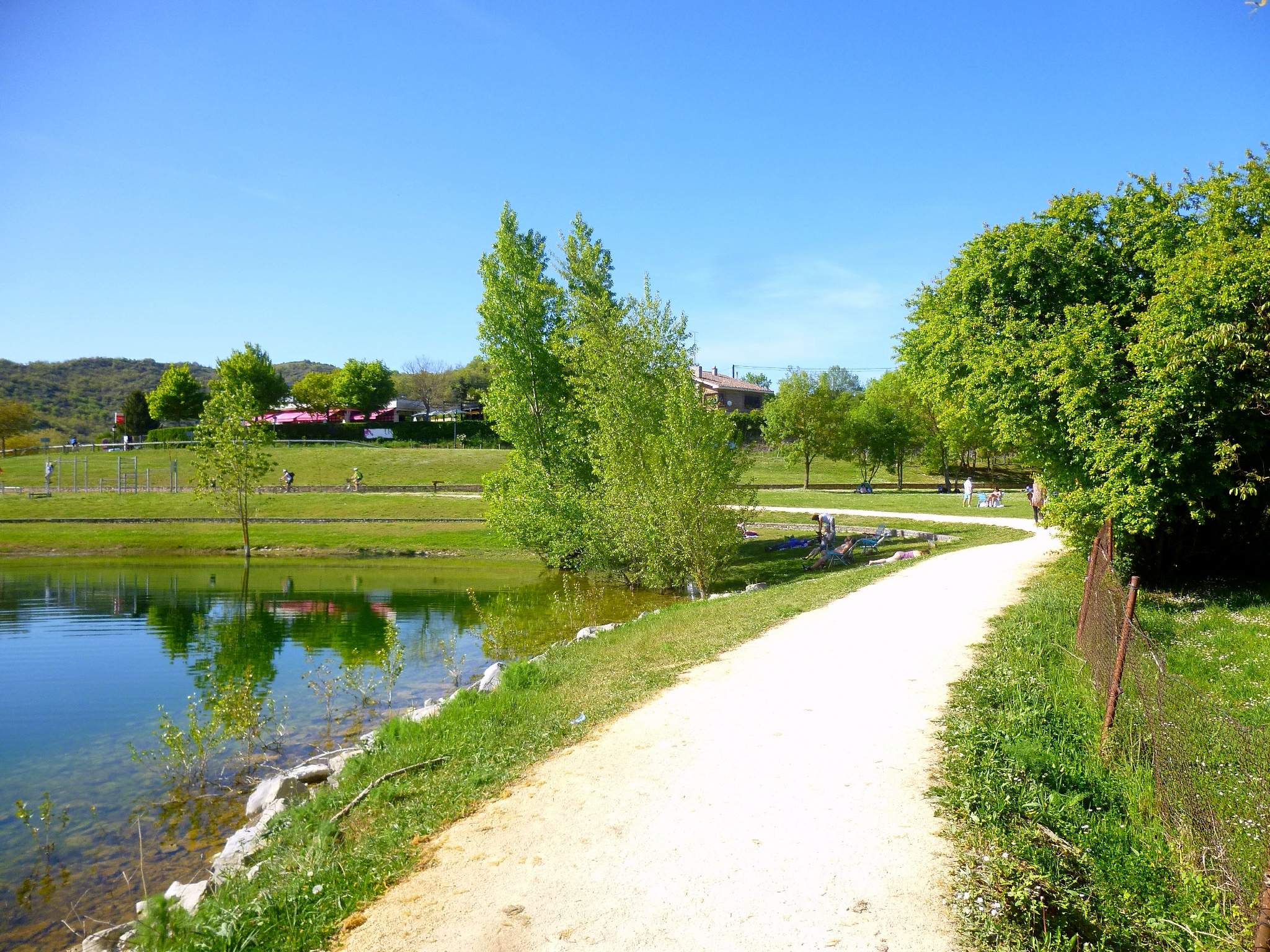 Ein Schotterweg neben einem Gewässer, umgeben von üppiger Vegetation und einem Zaun, mit Menschen, die die Landschaft genießen, Gebäuden, Hügeln und einem klaren blauen Himmel im Hintergrund, der einen idealen Rahmen für eine Fahrradtour in Frankreich bildet.