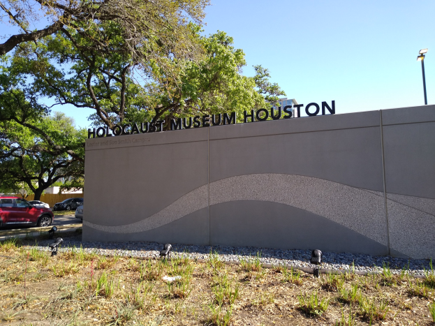 Außenansicht des Houston Holocaust Museums mit einer Gedenkwand, Steinen, Grünflächen, Bäumen, vorbeifahrenden Fahrzeugen, einem Straßenpfahl und einem bewölkten Himmel.