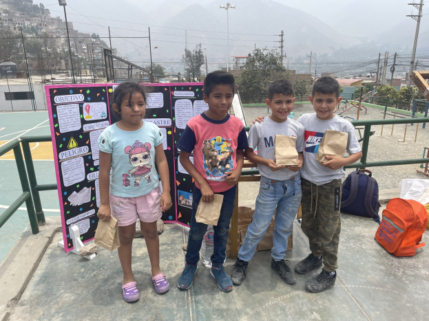 Eine Gruppe von Kindern hält Papiertüten in der Hand und steht auf einem Basketballfeld mit Tüten und Gegenständen rechts daneben und einer Tafel mit Text links daneben, mit Absperrungen, Pfosten und Gebäuden im Hintergrund.