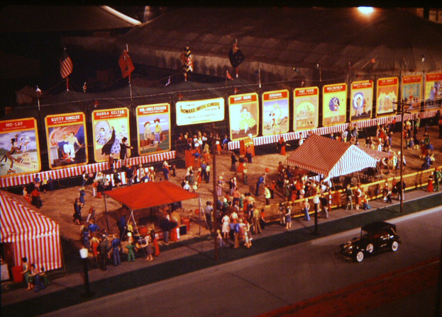 Ein altes Nachtfaschingsbild mit Menschen, einem Auto, Masten mit Fahnen, einem Zaun, einem Zelt, Brettern mit Bildern und Text und einer Straßenlaterne.