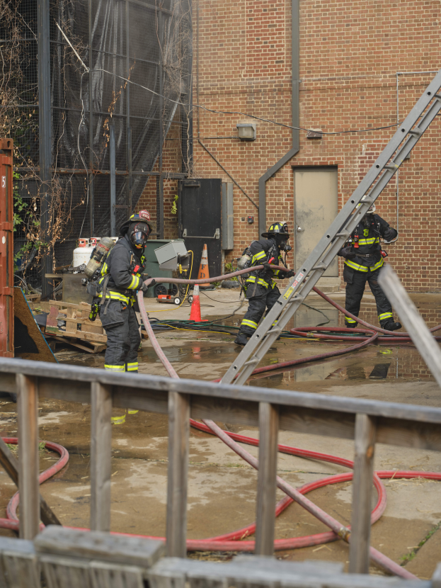 Eine Gruppe von Feuerwehrleuten in Helmen, die Rohre halten, arbeiten daran, ein Gebäude Feuer zu löschen, mit verschiedenen Ausrüstungen und Strukturen drumherum, einschließlich eines Metallzauns, Rohre auf dem Boden, einem Container, einem Verkehrskegel und einem Baum im Hintergrund.