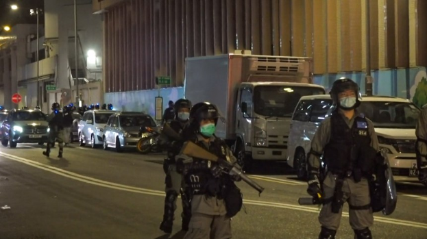 Eine Gruppe von Polizisten in Helmen, Handschuhen und Gesichtsmasken steht auf einer Straße bei Nacht, einige halten Waffen, mit Fahrzeugen, Laternenmasten, Schilderpfosten und Gebäuden im Hintergrund.