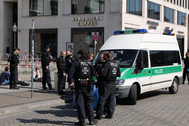 Eine Gruppe von Polizeibeamten vor einem Starbucks-Café, mit einem Van auf der rechten Seite und einigen Menschen auf der linken Seite, vor einer Hintergrundarchitektur aus einem Gebäude, einem Schild, einem Laternenmast und einem Zaun.