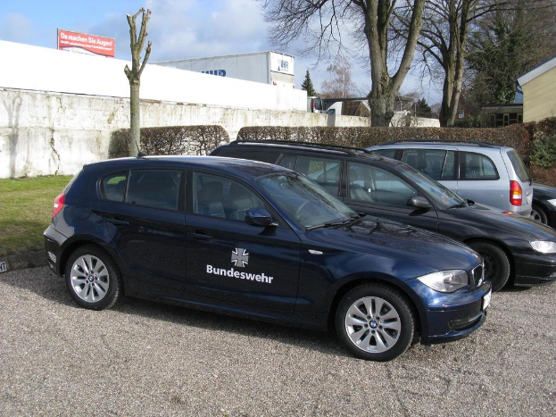 Mehrere Autos stehen auf einem Weg, der von grünem Gras, Büschen und Bäumen umgeben ist, mit einem Schild im Hintergrund, auf dem Text und ein Logo zu sehen sind, und Gebäuden in der Ferne.
