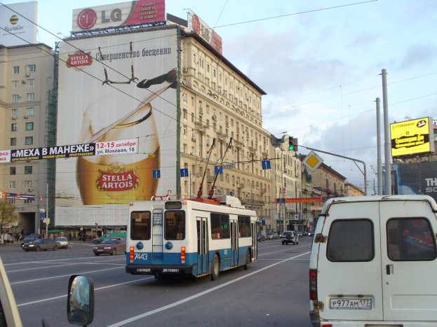 Eine belebte Stadtstraße mit Fahrzeugen, darunter ein Bus und ein Lieferwagen, umgeben von Gebäuden, Schildern, Strommasten mit Drähten und Verkehrsampeln unter einem bewölkten Himmel.