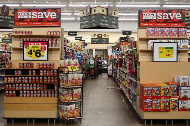 Ein Innenraum eines Lebensmittelladens mit verschiedenen auf Regalen, Gestellen und dem Boden ausgelegten Lebensmitteln, Deckenleuchten und Informationsschildern im Hintergrund.