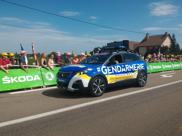 Ein Polizeiwagen fährt auf einer Straße neben einer Menschenmenge, einige tragen Mützen und halten Fahnen, mit einem Banner auf dem Wagen und einer Kulisse aus Bäumen, Gebäuden und einem bewölkten Himmel. Unten steht der Text "Gendarmerie de la Tour de France".