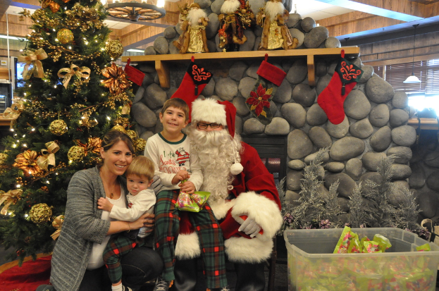 Eine Frau, Kinder und ein Mann in Santa-Klaus-Kostüm mit Brille und Handschuhen stehen neben einem geschmückten Weihnachtsbaum, einer Wand mit Steinen, Spielzeugen, Dekorationen, einem Korb mit Gegenständen und Hintergrundbeleuchtung, wobei ein Junge ein Päckchen hält.