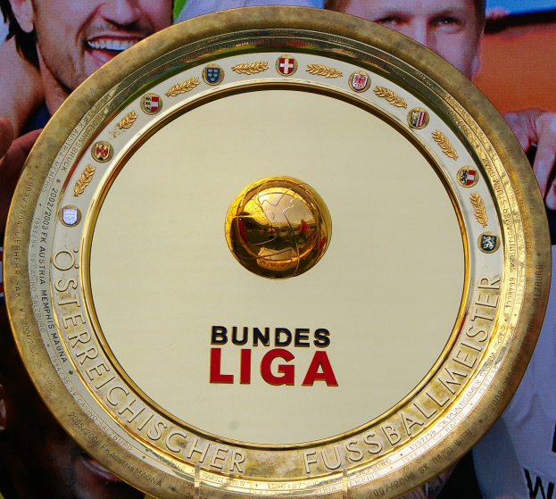 Goldener Schild mit 'Bundesliga'-Text in den Händen eines Mannes, mit einem Banner im Hintergrund, das drei Personen zeigt.