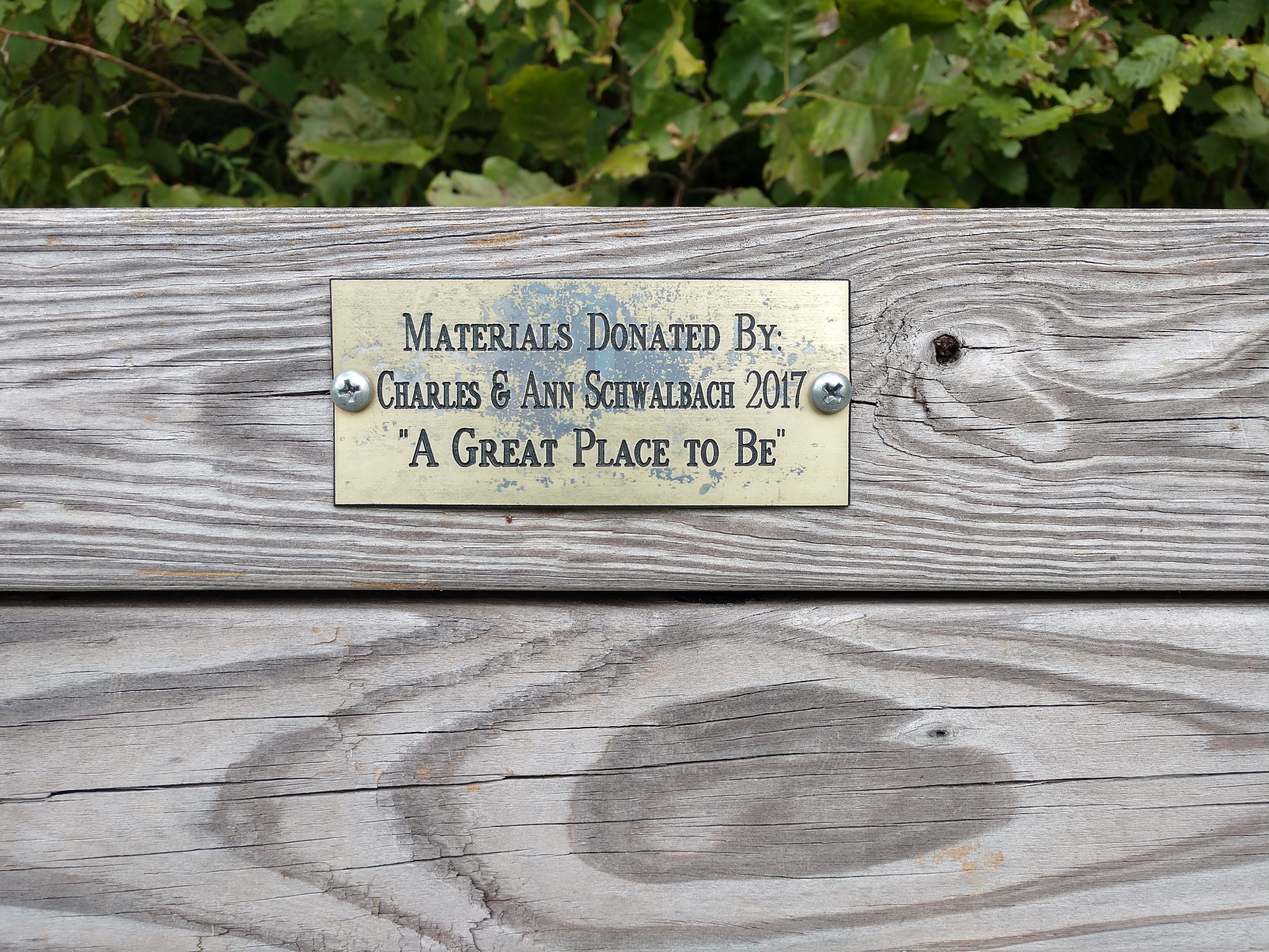 Holzbank mit einer Plakette, auf der "Materialien gespendet von Charles & Ann Schwabach 2017: Ein schöner Ort" steht, vor üppigen grünen Pflanzen.