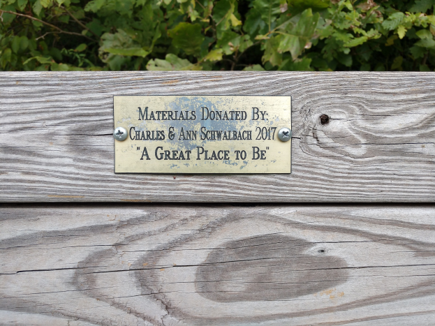 Holzbank mit einer Plakette, auf der "Materialien gespendet von Charles & Ann Schwabach 2017: Ein schöner Ort" steht, vor üppigen grünen Pflanzen.