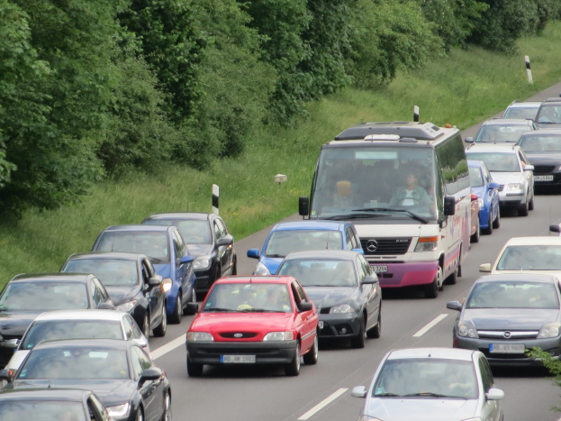 Ein Stau auf einer Autobahn mit vielen Autos und einem Lieferwagen, Menschen sind in den Fahrzeugen zu sehen, mit Bäumen und Gras im Hintergrund.