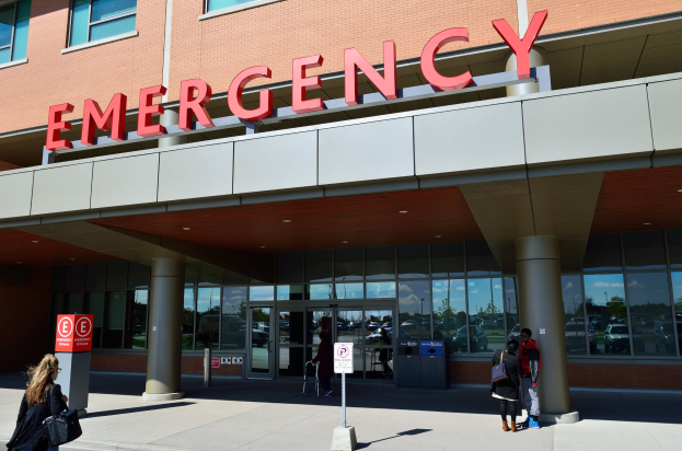 Eine Gruppe von Menschen geht vor einem Notaufnahme-Gebäude mit Glaswänden, Säulen, einem Namensschild, Hinweistafeln auf Pfählen und einem Müllbehälter.