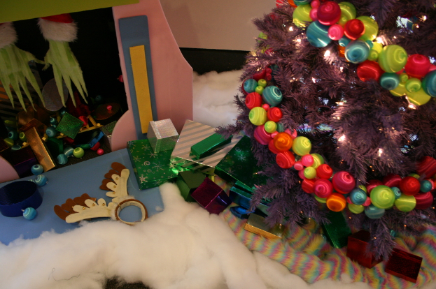 Drinnen steht ein geschmückter Weihnachtsbaum mit Lichterkette und Schmuck rechts, Geschenkboxen und Deko in der Mitte, eine Hütte mit Deko oben links und Baumwolle und Wolle unten.