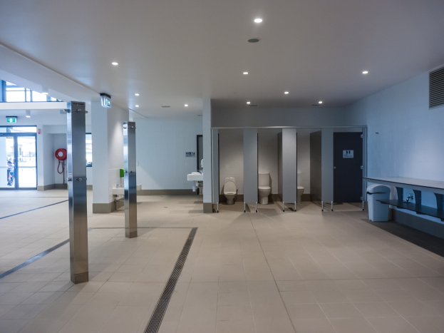 Große öffentliche Toilette mit mehreren Toiletten, tragenden Säulen, einem Mülleimer, einer Tür, einem Schild, einem an der Wand montierten Feuerlöscher und Deckenleuchten auf dem Dach.