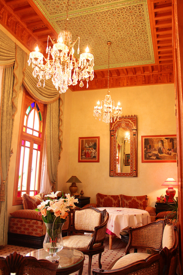 Ein Wohnzimmer mit einem Tisch und Blumentopf im Vordergrund, umgeben von Stühlen, Sofas und einem Tisch mit einer weißen Decke im Hintergrund, gerahmte Bilder an der Wand, von der Decke hängende Lampen und Vorhänge mit einem Fenster auf der linken Seite.