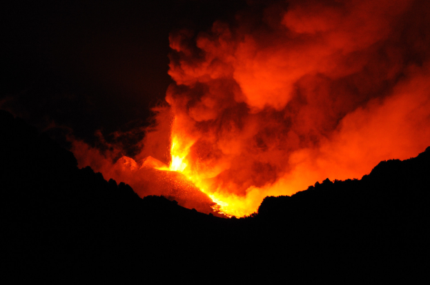 Ein Vulkanausbruch bei Nacht, mit leuchtend orangefarbenem und gelbem Lavafluss ins Meer und Rauch, der in den dunklen Himmel aufsteigt.