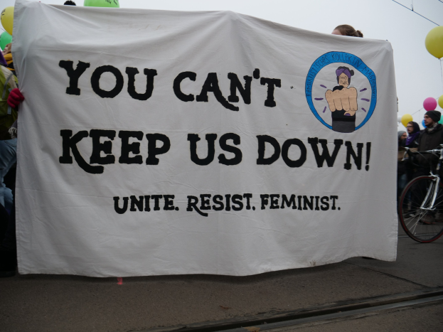 Gruppe von Menschen hält ein 'Du kannst uns nicht unterdrücken, vereine widerstehe Feminist' -Schild auf einer Straße mit einem Fahrrad in der Nähe, Ballons und Drähte im Hintergrund unter einem klaren blauen Himmel.