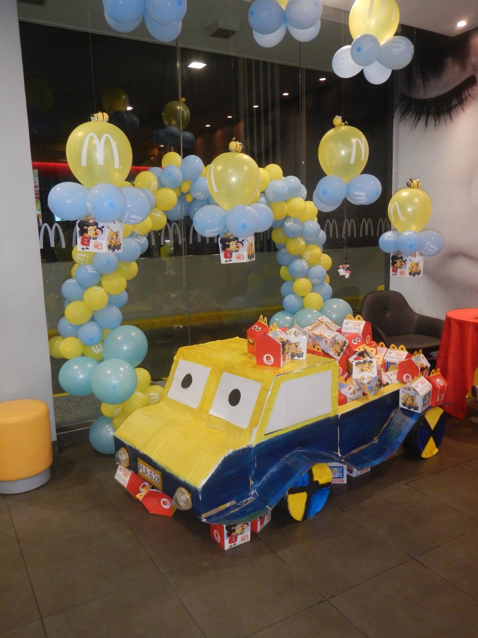 Eine festliche McDonald's-Geburtstagsparty-Szene mit einem gelben und blauen dekorierten Truck mit Ballons, einem rot gedeckten Tisch mit Gegenständen, Hockern, einer Glaswand und Deckenlampen.