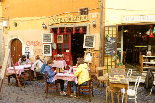 Eine Gruppe von Menschen an Tischen eines Restaurants in Rom, Italien, mit Flaschen, Gläsern und Dekoration auf den Tischen und beleuchteten Wänden mit Text und Regalen mit Gegenständen im Inneren.