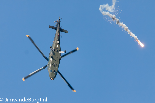 Ein militärisches Helikopter in der Luft, das eine Rakete hält, die Rauch und Feuer ausstößt, mit Text in der unteren linken Ecke.