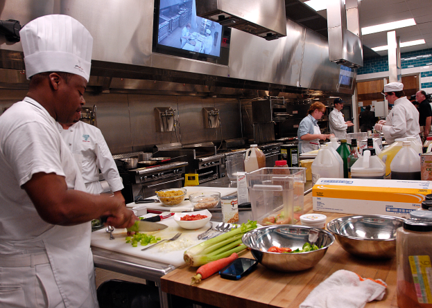 Eine Gruppe von Köchen in einer Küche beim Zubereiten von Speisen auf einem Schneidbrett, wobei ein Koch im Vordergrund in einer weißen Schürze und Mütze steht und ein Messer hält und von verschiedenen Utensilien, Schüsseln, Gemüse, einem Mobiltelefon, Flaschen und anderen Gegenständen umgeben ist, mit einem Fernseher und Deckenleuchten im Hintergrund.