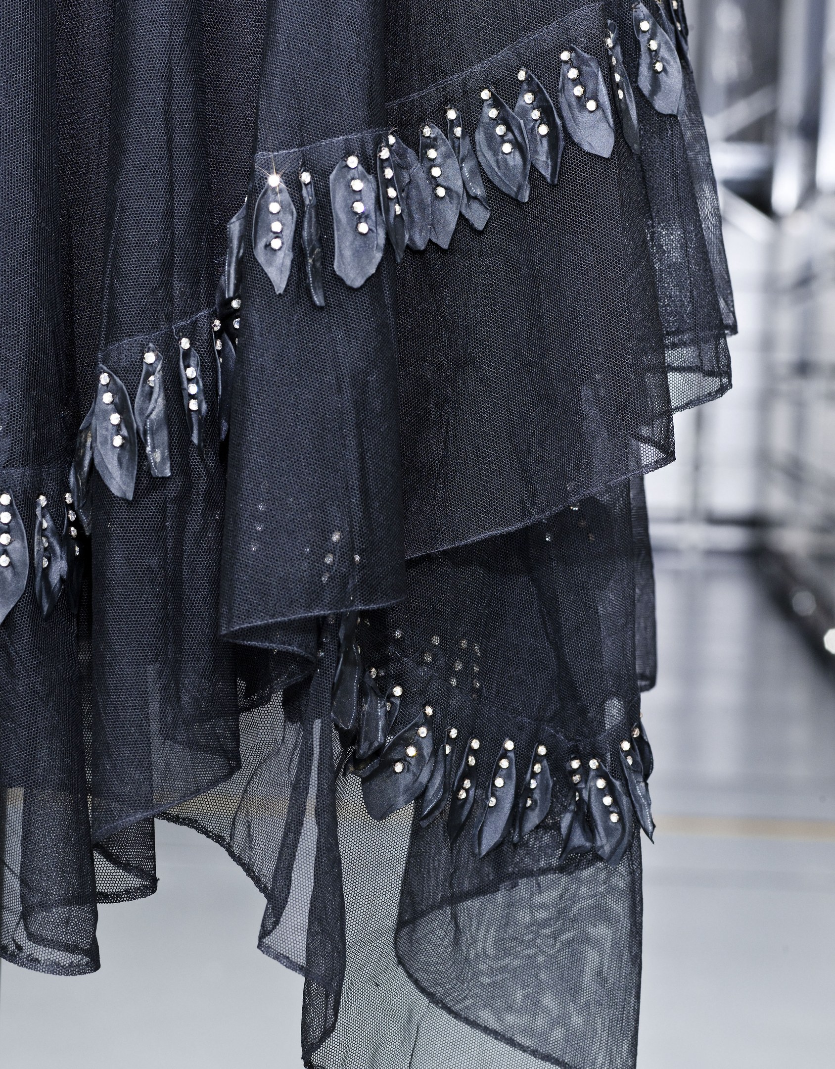 Schwarzes bodenlanges Kleid mit hohem Ausschnitt, langen Ärmeln und vollem Rock, mit aufwendiger Perlen- und Paillettenverzierung auf leichtem Stoff mit feiner Schimmer, präsentiert auf dem Laufsteg.