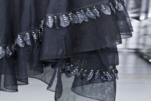 Schwarzes bodenlanges Kleid mit hohem Ausschnitt, langen Ärmeln und vollem Rock, mit aufwendiger Perlen- und Paillettenverzierung auf leichtem Stoff mit feiner Schimmer, präsentiert auf dem Laufsteg.