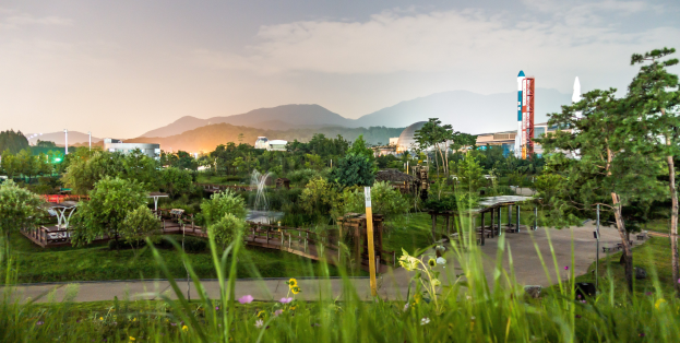 Ein Park mit grünem Gras, bunten Blumen, hohen Bäumen, einer Brücke, einem Springbrunnen, Laternenmasten und Gebäuden im Hintergrund, mit Bergen und einem klaren blauen Himmel.