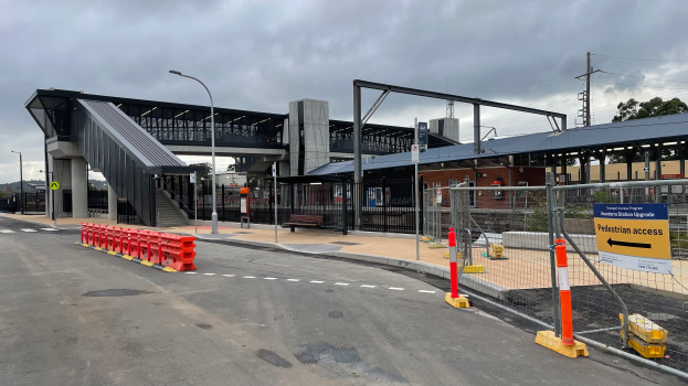Ein Bahnhofsgebäude mit einer Brücke, Straßenlaternen, Strommasten, Stromkabeln, Bäumen, Gebäuden, Kraftfahrzeugen, Absperrgittern, Schildern, Bänken und einer bewölkten Himmel.