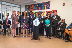 Gruppe von Menschen in einem Raum mit einem Podium, Mikrofon und einer Tafel, die "Sancho City Councilman Joins City Council" anzeigt, umgeben von Flaggen, einem Feuerlöscher und Deckenleuchten.