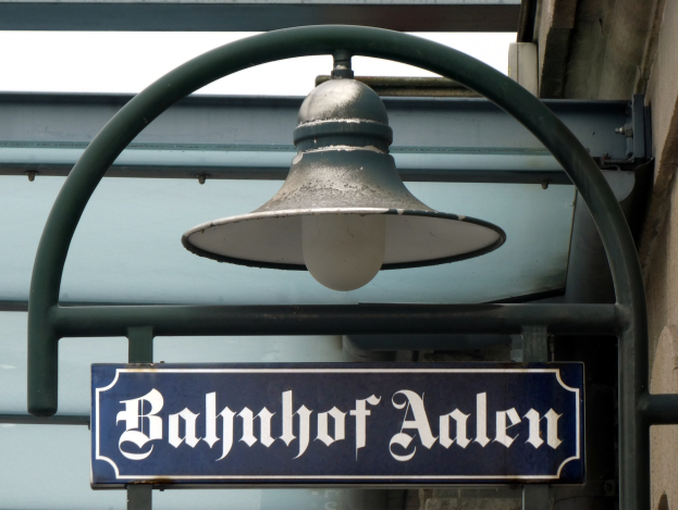 Ein Schild mit der Aufschrift "Bahnhof Aalen" hängt an einem Gebäude, beleuchtet von einer wandmontierten Lampe, mit einer Glasdecke im Hintergrund.