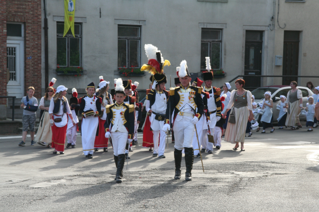 Eine Gruppe von Menschen in Kostümen und mit Stöcken marschiert auf einer Straße in einem Umzug, mit Gebäuden, Pflanzen, einer Fahne und Fahrzeugen im Hintergrund.