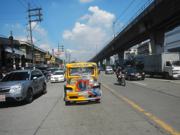 Gelbes Taxi fährt auf einer Straße mit hohen Gebäuden, Strommasten mit Drähten und einer Brücke im Hintergrund unter einem bewölkten Himmel, mit einem Wasserzeichen auf dem Bild.