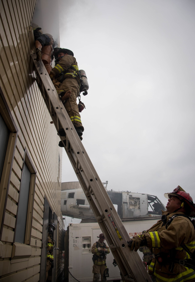 Feuerwehrleute in Helmen, die ein Gebäude mit einer Leiter erklimmen, mit einem Fahrzeug und Himmel im Hintergrund.