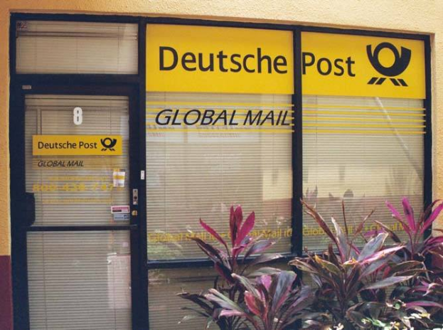 Ein Geb├Ąude mit einer T├╝r, Jalousien und Pflanzen davor, mit einem Schild, auf dem "Deutsche Post Global Mail" steht.