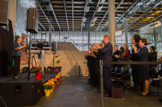 Eine Gruppe von Menschen auf der Bühne, die klatschen, während eine Frau ein Mikrofon hält; Blumentöpfe, ein Lautsprecher und Tische mit Gegenständen sind im Hintergrund zu sehen, mit einer Menschenmenge, Stühlen, einer Treppe, Säulen, Deckenleuchten und Fenstern.