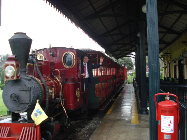 Ein alter roter Zug mit einem Mann in einem schwarzen Anzug, der an der Tür steht und lächelt und posiert, mit einer Stationshütte auf der rechten Seite und einem roten Feuerlöscher im Vordergrund.
