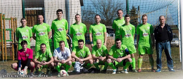 Eine Gruppe von Männern in grünen T-Shirts steht und hockt auf einem Fußballfeld mit einem Ball davor, einem Torpfosten rechts daneben und einem Hintergrund aus Bäumen, Gebäuden, Pfählen, Fahrzeugen und dem Himmel, mit Text unten links.