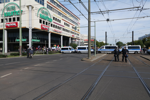 Eine Gruppe von Polizisten steht an der Straßenseite neben einer Tram, mit Fahrzeugen, Strommasten, Gebäuden mit Schildern, Bäumen und einem bewölktem Himmel im Hintergrund.
