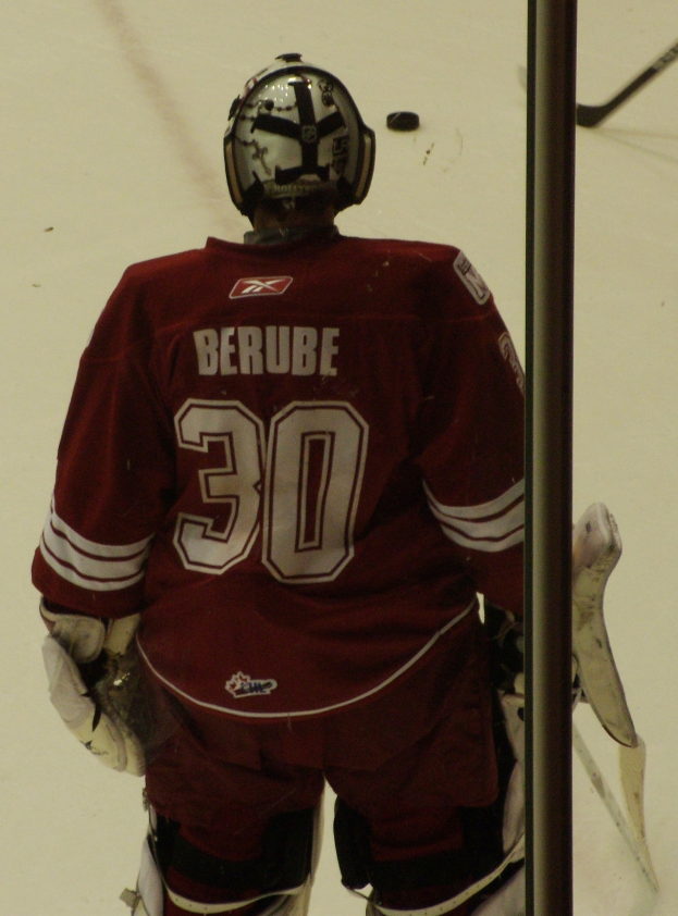 Ein Hockey-Spieler in Helm, Handschuhen und einem nummerierten Trikot steht auf dem Eis vor einem Tor mit einem Pfosten rechts daneben.
