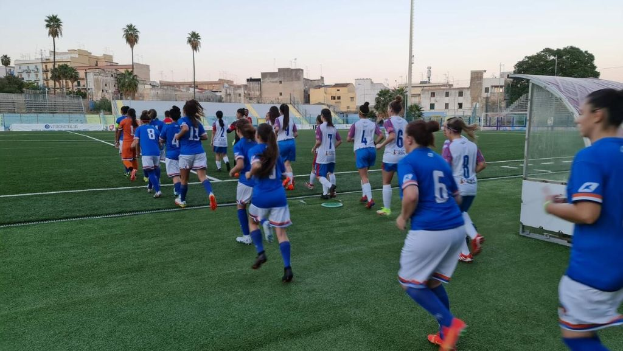 Eine Gruppe von Frauen, die auf einem Feld mit Gebäuden, Bäumen und Pfählen umgeben unter einem klaren blauen Himmel Fußball spielen, mit einem Torpfosten und Netz auf der rechten Seite.