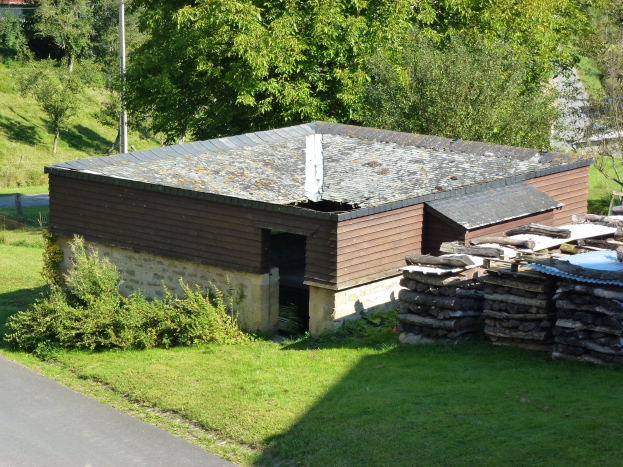 Kleines Holzhaus neben einer Straße, umgeben von Gras, Pflanzen, Bäumen und einem Pfahl, mit gestapelten Holzscheiten rechts.