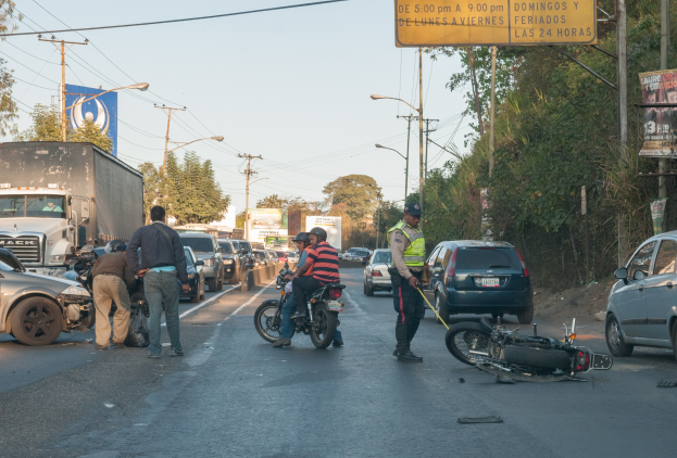 Eine Gruppe von Menschen steht um ein verunglücktes Motorrad am Rande einer Straße mit mehreren Fahrzeugen, darunter ein Lastwagen, und einer Hintergrund von Bäumen, Masten, Lichtern und Schildern unter dem Himmel.
