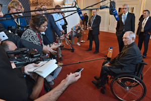 Ein Mann im Rollstuhl umgeben von Reportern mit Kameras, Mikrofonen und Handys sowie einem Banner und Lichtern im Hintergrund.